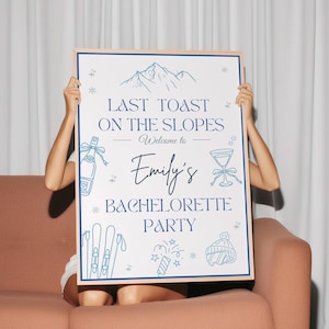 May include: A framed sign with a blue border and text that reads "Last Toast on the Slopes, Welcome to Emily's Bachelorette Party." The sign features illustrations of mountains, skis, and cocktail glasses. The sign is held up by a person sitting on a pink couch.