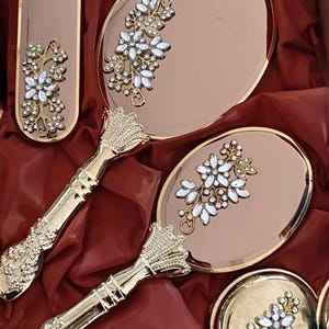 Op de afbeelding: Een set roségouden kaptafelaccessoires bestaande uit twee handspiegels, een langwerpig rechthoekig dienblad en een ronde houder. Elk item heeft een metallic afwerking en is versierd met een cluster van heldere, gefacetteerde bloemkristallen.