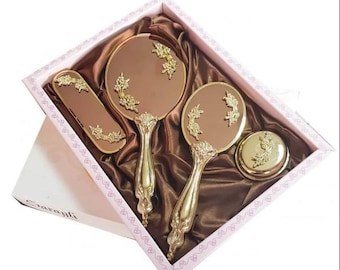 May include: A vintage vanity set in a pink box. The set includes two hand mirrors, a brush, and a compact, all with gold-colored ornate detailing. The mirrors and brush have brown surfaces. The set is presented on a brown satin fabric.
