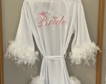 May include: White satin robe with a pink embroidered crown and diamond design and the word "Bride". The robe has white feather trim on the sleeves and hem.