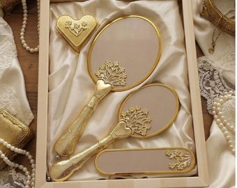 May include: A set of vintage-style vanity accessories, including two hand mirrors, a small rectangular mirror, and a heart-shaped compact, all with gold-colored frames and handles. The set is presented in a wooden box lined with satin.