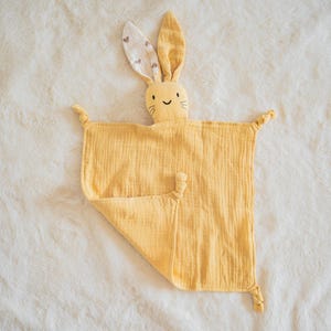 以下が含まれることがあります： ウサギの頭が付いた黄色のベビー用セキュリティブランケット。ウサギはハート模様の長い耳と笑顔の顔をしています。ブランケットは柔らかく、質感のある生地で作られています。