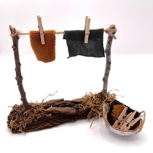 K&ouml;nnte beinhalten: Eine Miniatur-W&auml;scheleine aus Zweigen und Schnur, mit zwei kleinen Stoffquadraten, die mit Holzklammern an der Leine befestigt sind. Ein kleiner Korb enth&auml;lt zus&auml;tzliche W&auml;scheklammern und Stoffst&uuml;cke. Die Basis besteht aus Rinde und Moos.