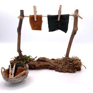 K&ouml;nnte beinhalten: Miniatur-W&auml;scheleine im rustikalen Stil mit zwei kleinen Stoffquadraten, die mit Holzklammern befestigt sind. Die W&auml;scheleine wird von zwei Holzpfosten getragen und steht auf einem Sockel aus Holz und Moos. Ein kleiner Korb enth&auml;lt zus&auml;tzliche W&auml;scheklammern und Stoff.