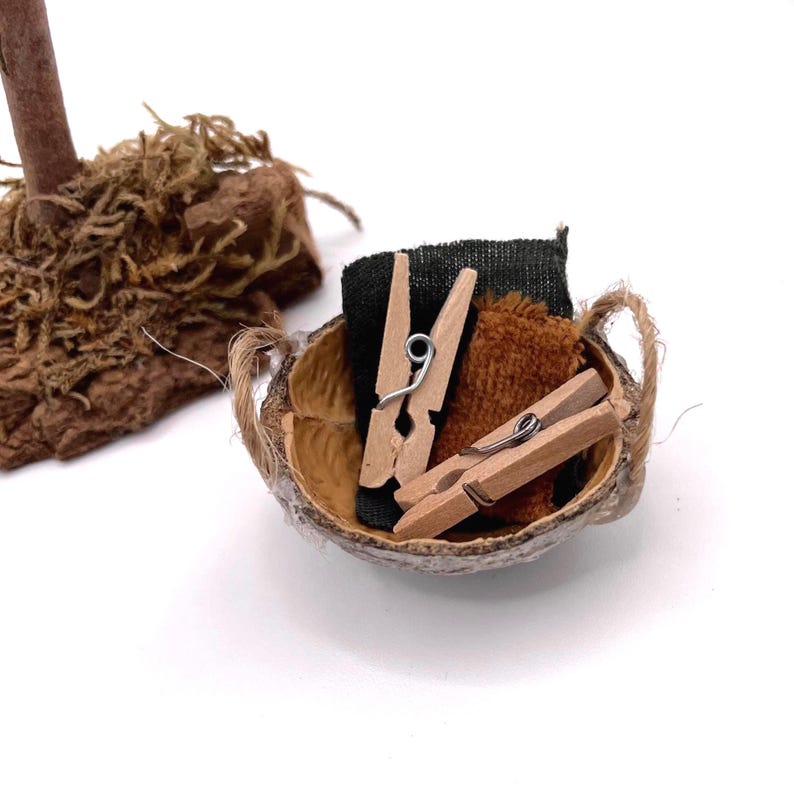 K&ouml;nnte beinhalten: Ein Miniaturkorb aus Kokosnussschale, gef&uuml;llt mit kleinen W&auml;scheklammern aus Holz und Stoffresten. Der Korb hat einen geflochtenen Griff und ist vor einem wei&szlig;en Hintergrund platziert. Die Stoffreste sind schwarz und braun.