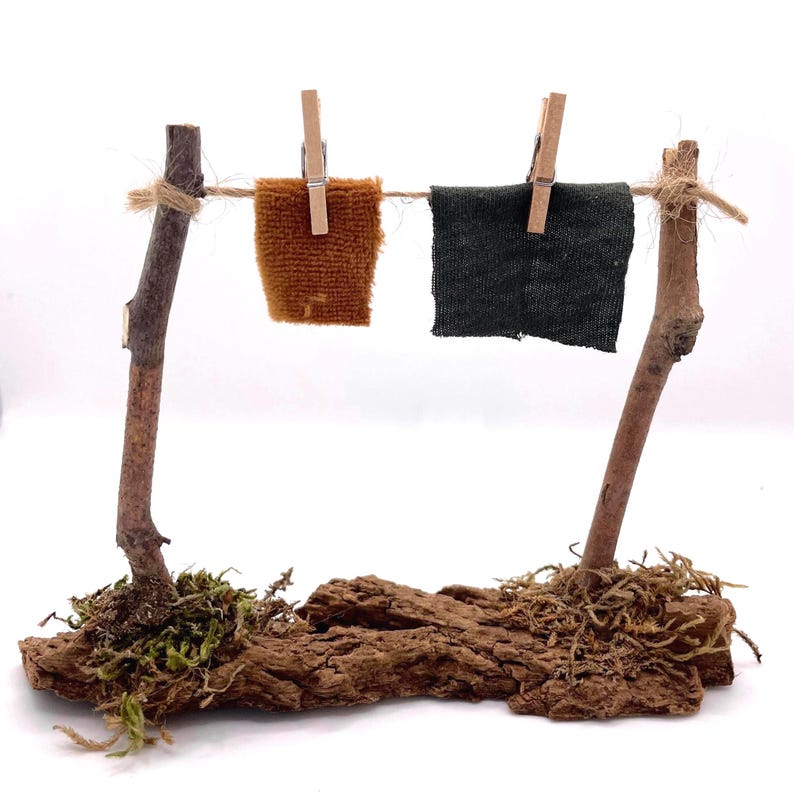 K&ouml;nnte beinhalten: Miniatur-W&auml;scheleine mit zwei kleinen Stoffquadraten, eines braun und eines dunkelgr&uuml;n, mit Holzklammern befestigt. Die W&auml;scheleine wird von zwei Holzpfosten getragen, die an einer Rindenbasis mit Moos befestigt sind.