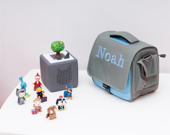 Könnte beinhalten: Eine graue und blaue personalisierte Tragetasche mit dem Namen "Noah", der auf der Vorderseite gestickt ist. Die Tasche steht auf einem weißen Tisch mit verschiedenen kleinen Spielzeugen, darunter ein Pinguin, ein Wolf und ein Baum.