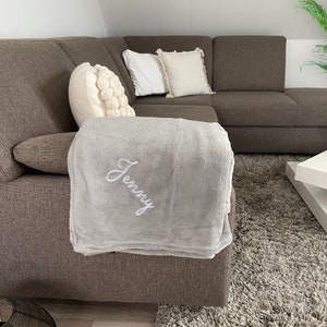 Op de afbeelding: Een grijze deken met de naam "Jenny" geborduurd in wit op een grijze bank.