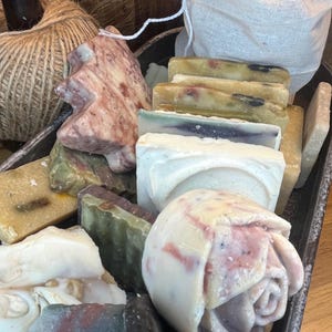 Può includere: Una collezione di saponi fatti a mano in varie forme e colori. I saponi includono un sapone a forma di albero, un sapone a forma di rosa e barre rettangolari con diversi strati di colore. I saponi sono disposti in un vassoio di metallo.