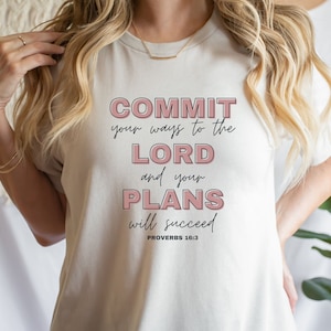 May include: Cream-colored t-shirt with the text "COMMIT your ways to the LORD and your PLANS will succeed" in pink and black lettering. The text is from Proverbs 16:3. The shirt has a crew neck.