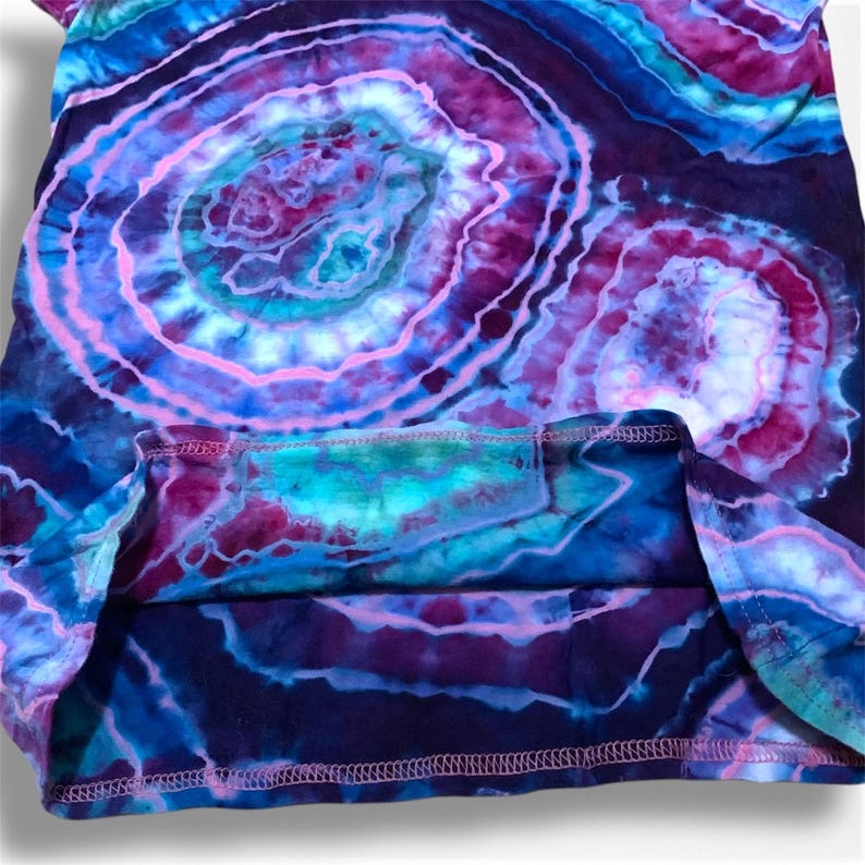 May include: A tie-dye shirt with a purple, blue, and pink swirl pattern. The shirt has a hemmed edge and a relaxed fit.