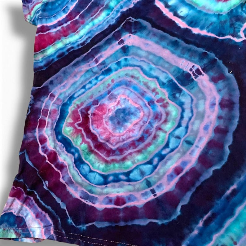 May include: A tie-dye shirt with a circular pattern in shades of blue, pink, purple, and green. The shirt has a dark blue background with a large, central circle.