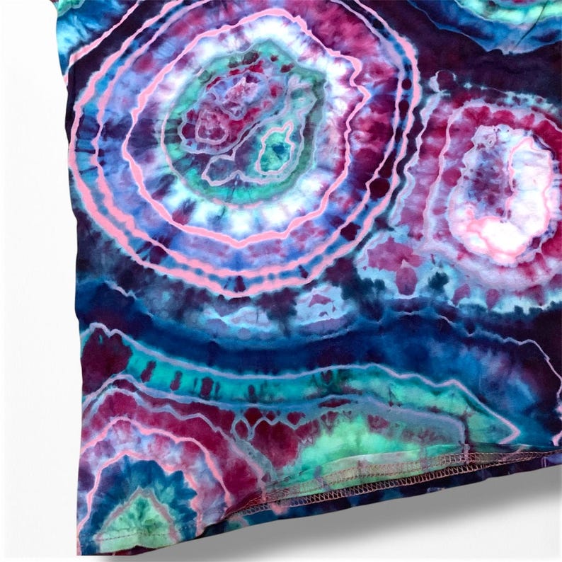 May include: A close-up of a tie-dye fabric with a swirling pattern of pink, blue, and green colors. The fabric has a textured surface and a subtle sheen.