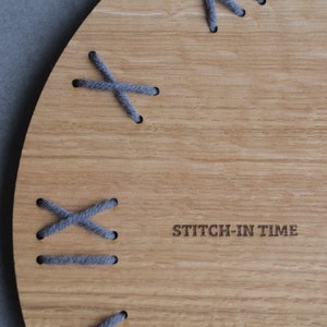 May include: A wooden clock face with gray stitching around the edge. The clock face has the text "STITCH-IN TIME" printed on it.