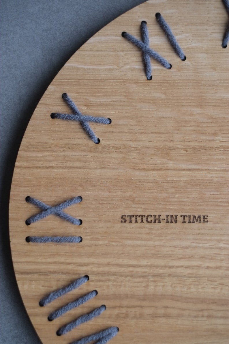 May include: A wooden clock face with gray stitching around the edge. The clock face has the text "STITCH-IN TIME" printed on it.