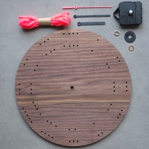 May include: A wooden clock face with holes for stitching, a pink yarn ball, two black clock hands, a pink clock hand, a clock mechanism, two metal washers, and a gold metal washer. The clock face has the text "Stitch in Time" printed on it.