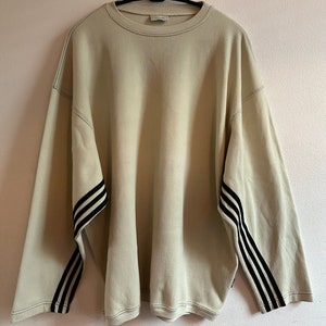 May include: Oversized beige sweatshirt with black stripes on the sleeves. The sweatshirt has a crew neck and long sleeves. The Adidas logo is visible on the hanger. Stitching is visible around the neckline and sleeves.