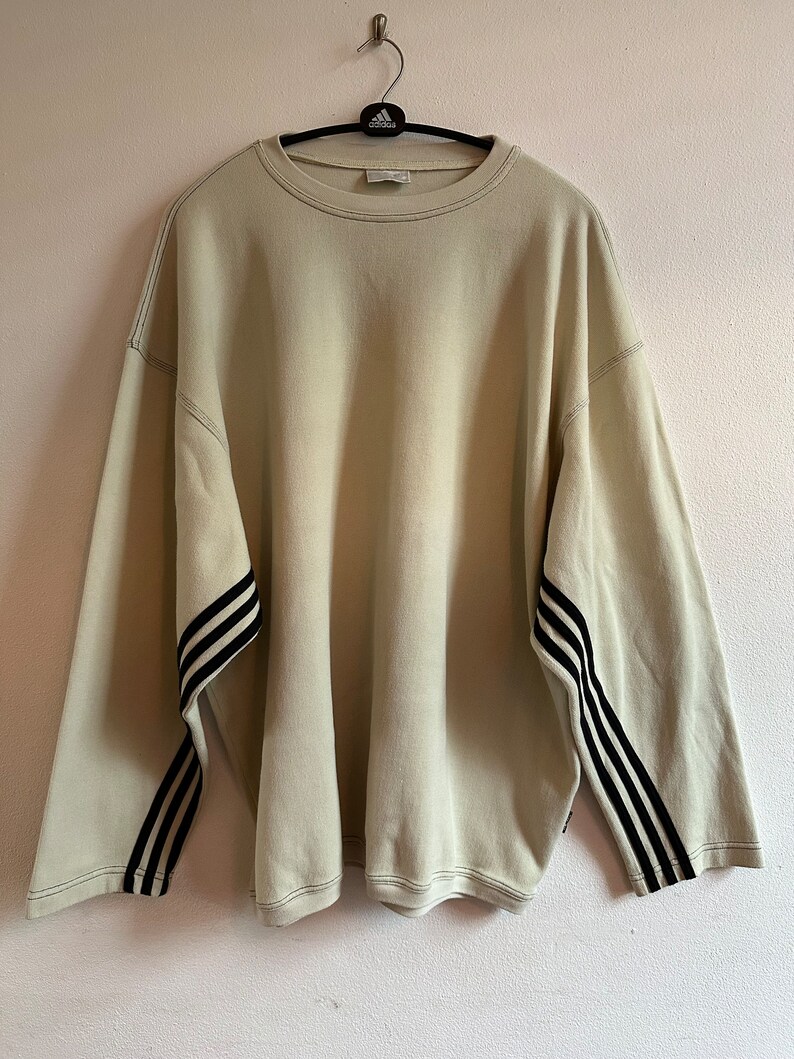 May include: Oversized beige sweatshirt with black stripes on the sleeves. The sweatshirt has a crew neck and long sleeves. The Adidas logo is visible on the hanger. Stitching is visible around the neckline and sleeves.