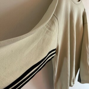 May include: Oversized beige sweatshirt with black stripes on the sleeves. The long-sleeved top is hanging on a black hanger against a light-coloured wall. The sweatshirt has a crew neck and a relaxed fit.