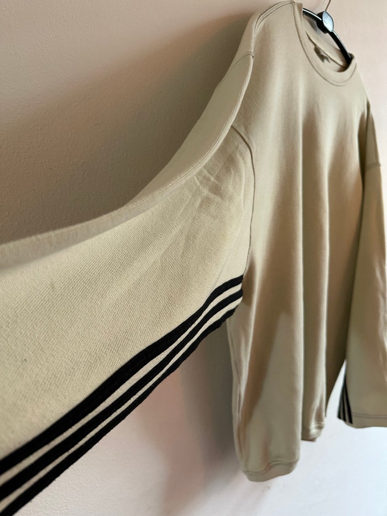 May include: Oversized beige sweatshirt with black stripes on the sleeves. The long-sleeved top is hanging on a black hanger against a light-coloured wall. The sweatshirt has a crew neck and a relaxed fit.