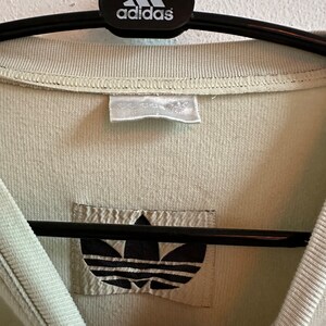 May include: Close-up of a pale green Adidas shirt. The shirt is hanging on a black hanger with the Adidas logo. The Adidas logo is also visible on a patch on the shirt.