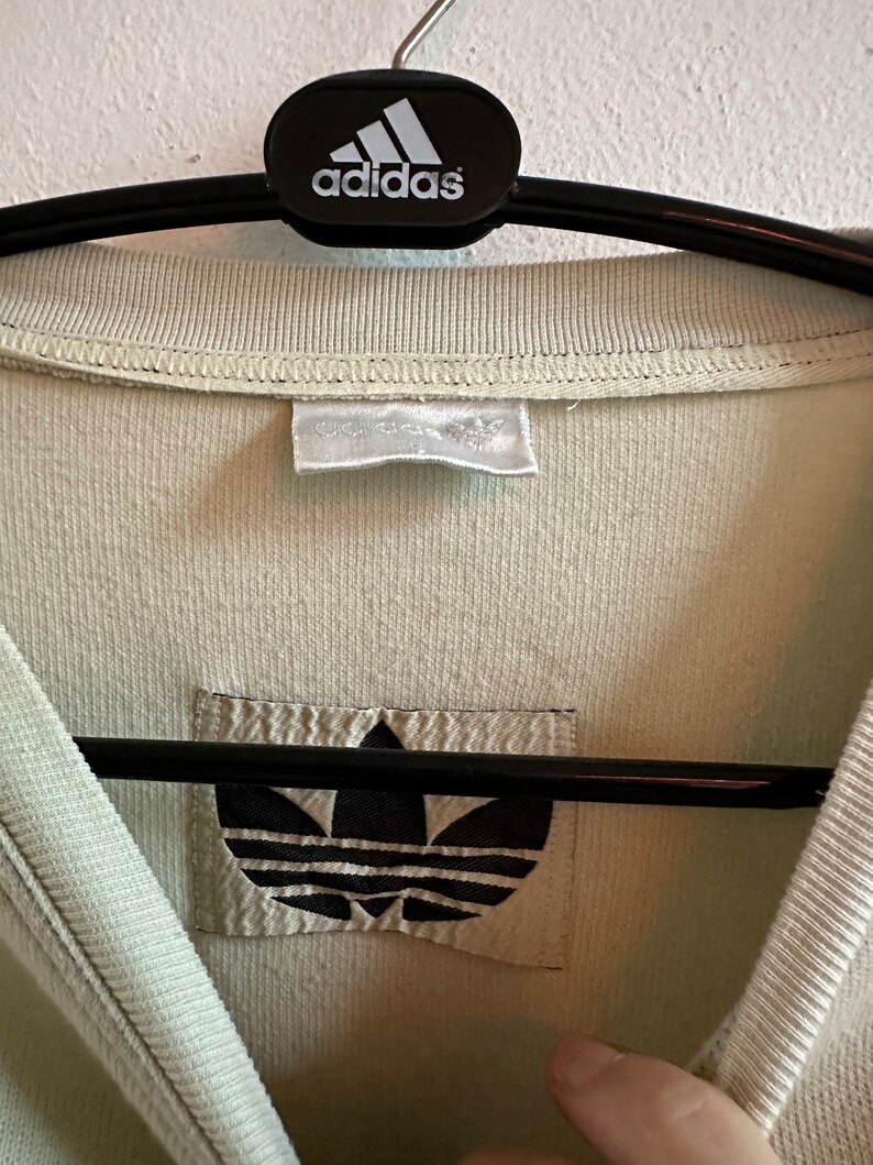 May include: Close-up of a pale green Adidas shirt. The shirt is hanging on a black hanger with the Adidas logo. The Adidas logo is also visible on a patch on the shirt.