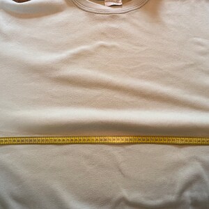 May include: A pale beige sweatshirt with a crew neck and long sleeves. A yellow measuring tape is stretched across the chest of the sweatshirt, indicating a measurement. The sweatshirt has a simple, classic design.