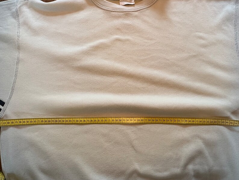 May include: A pale beige sweatshirt with a crew neck and long sleeves. A yellow measuring tape is stretched across the chest of the sweatshirt, indicating a measurement. The sweatshirt has a simple, classic design.