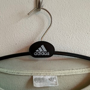 May include: A pale green Adidas t-shirt hanging on a black hanger. The Adidas logo is displayed on the hanger and the shirt's label. The t-shirt is made of a soft fabric and has a round neck.