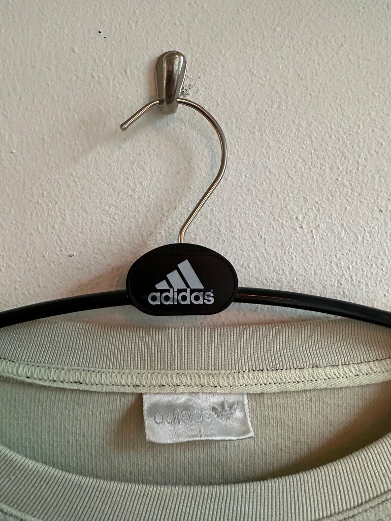May include: A pale green Adidas t-shirt hanging on a black hanger. The Adidas logo is displayed on the hanger and the shirt's label. The t-shirt is made of a soft fabric and has a round neck.