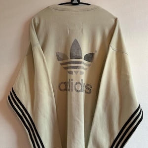 May include: A light beige Adidas sweatshirt featuring the Adidas logo and text on the back. The sleeves have black stripes. The sweatshirt is hanging on a hanger.