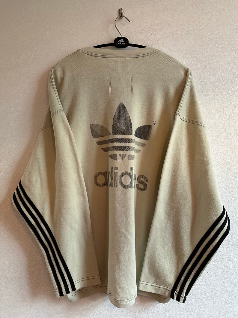 May include: A light beige Adidas sweatshirt featuring the Adidas logo and text on the back. The sleeves have black stripes. The sweatshirt is hanging on a hanger.