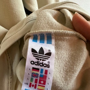 May include: Close-up of a beige Adidas sweatshirt label. The label features the Adidas logo, the word "adidas", and various international flags. The sweatshirt is a light beige colour.