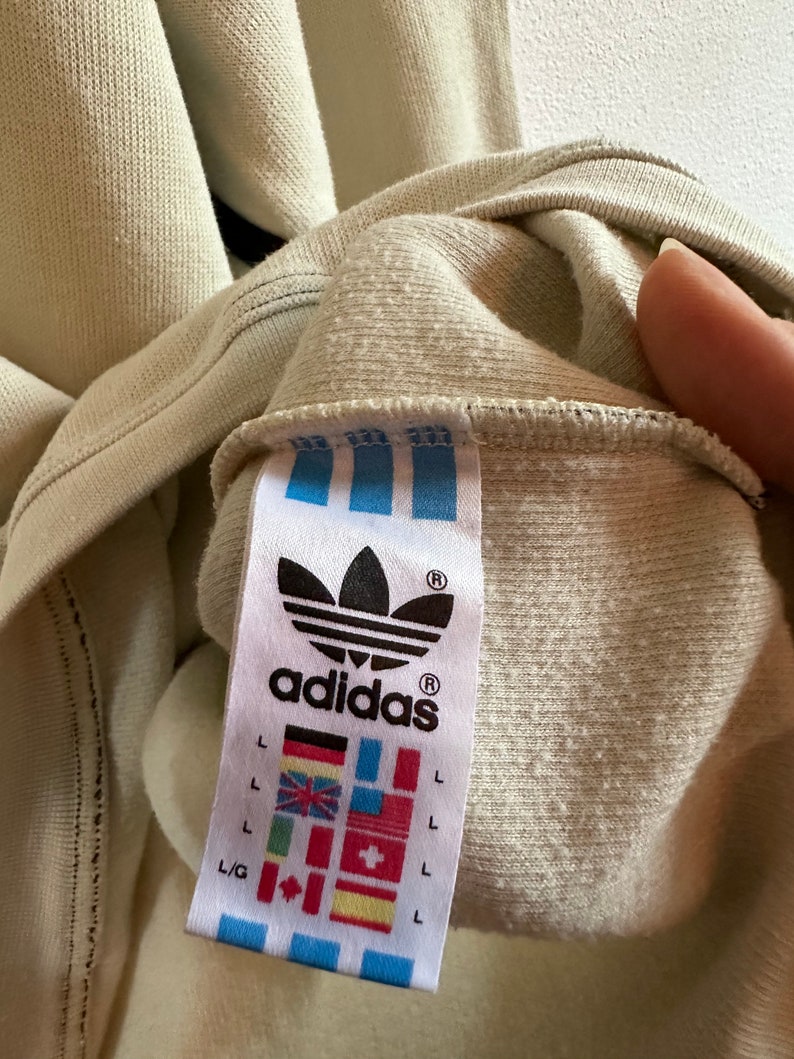 May include: Close-up of a beige Adidas sweatshirt label. The label features the Adidas logo, the word "adidas", and various international flags. The sweatshirt is a light beige colour.