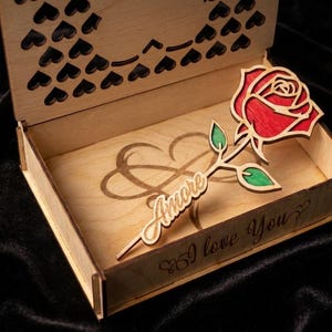Puede incluir: Caja de madera con una rosa cortada con láser y la palabra "Amore". La rosa es roja con hojas verdes. La caja tiene un diseño de corazón y las palabras "I love You" grabadas. Hecha de madera clara.