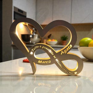 Può includere: Una luce a forma di cuore in legno con un simbolo dell'infinito. La luce è incisa con i nomi "Marco" e "Agata", e la frase italiana "Tutto è possibile se stiamo insieme". La luce è illuminata con un bagliore caldo.