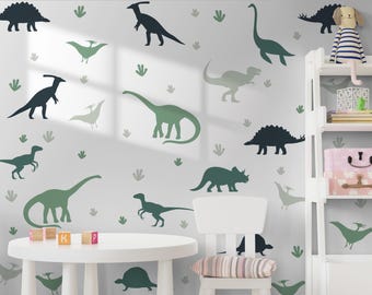 May include: A white wall adorned with dinosaur silhouettes in green and teal hues. A white table with wooden blocks and a white chair are positioned in front of the wall. A white shelf displays a stuffed dog and decorative items, creating a playful scene.