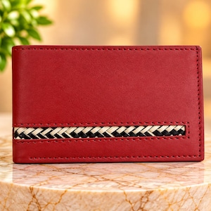 May include: A rectangular red leather wallet featuring a horizontal inset strip of woven black, white, and tan material across the lower section. The wallet has visible stitching along the edges and a smooth, matte finish on the red surface.
