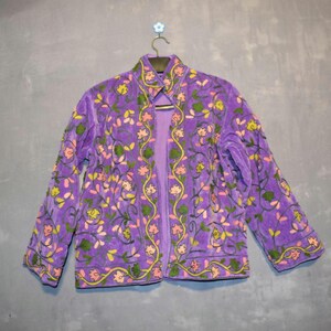 Peut inclure: Veste violette avec broderies florales. La veste présente une texture velours et est ornée de motifs floraux verts, jaunes et roses. La veste est suspendue à un cintre sur fond gris.