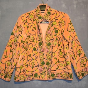 Peut inclure: Veste orange avec broderie florale. Le col est mandarin et les manches longues. La broderie comprend des fleurs et des vignes vertes, jaunes et roses. La veste est suspendue à un cintre sur fond bleu-gris.