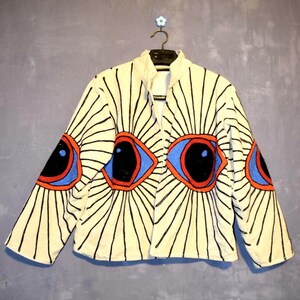 May include: Cream-coloured jacket with a unique eye design. The jacket features large, stylised eyes in red, blue, and black, with radiating black lines. The jacket is hanging on a black hanger against a grey wall.