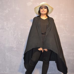 May include: A woman wearing a black cape and black pants stands in front of a light gray wall. She is wearing a white hat with a wide brim.