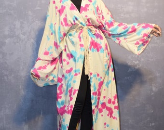 May include: A long, flowing tie-dye kimono with pink, blue, and white patterns. The kimono is open in the front and tied at the waist with a white sash. The model is wearing black pants and brown cowboy hat.