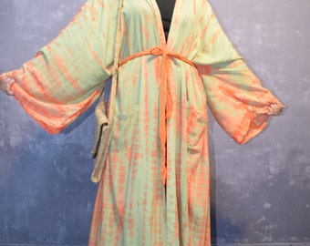 May include: A long, flowing robe with a tie-dye pattern in shades of green and orange. The robe has wide sleeves and pockets. A matching orange belt cinches the waist. The person is wearing a black hat and carrying a woven bag.