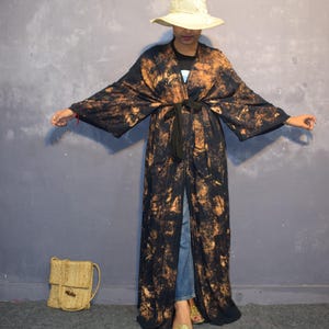 May include: A long, open-front kimono-style robe in black with a mottled orange pattern. The robe is tied at the waist with a black belt. A straw hat and a woven handbag are also visible.