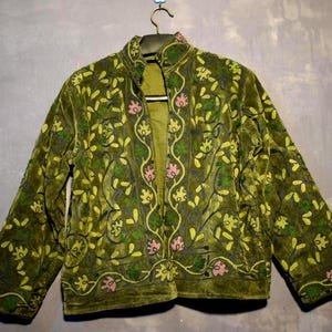Peut inclure: Veste en velours vert olive avec broderies florales jaunes, roses et vertes. La veste a un col mandarin et des manches longues. La broderie crée un motif détaillé sur la surface de la veste, ajoutant de la texture et de l'intérêt visuel.