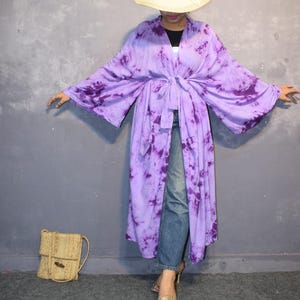 May include: A long, flowing, tie-dye kimono in shades of purple and violet. The kimono is tied at the waist and worn with blue jeans and gold heels. A straw hat and a woven bag complete the look. The background is a solid gray.