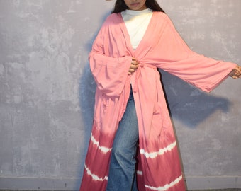 Puede incluir: Una larga bata de color rosa y blanco con estampado tie-dye, un sombrero blanco y unos vaqueros azules.