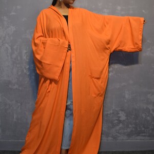 Może przedstawiać: Długi, pomarańczowy kimono o szerokim, swobodnym kroju. Kimono ma kaptur i długie rękawy z kieszenią po lewej stronie.
