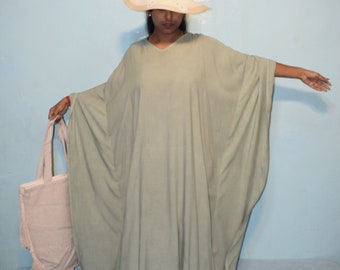 Op de afbeelding: Een lichtgroene kaftan jurk met een V-hals en wijde mouwen. De persoon draagt een beige zonnehoed en een lichtroze draagtas. Zwarte ballerina's maken de outfit compleet. De achtergrond is een lichtblauwe muur.